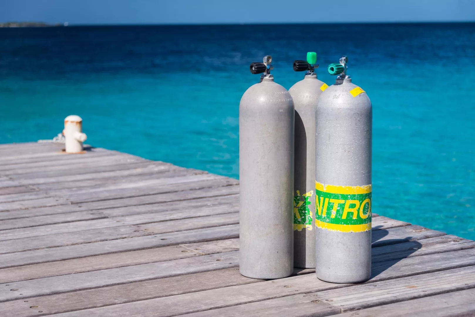 Three grey Nitrox scuba tanks with green and yellow labels on a wooden pier by clear blue tropical ocean water.
